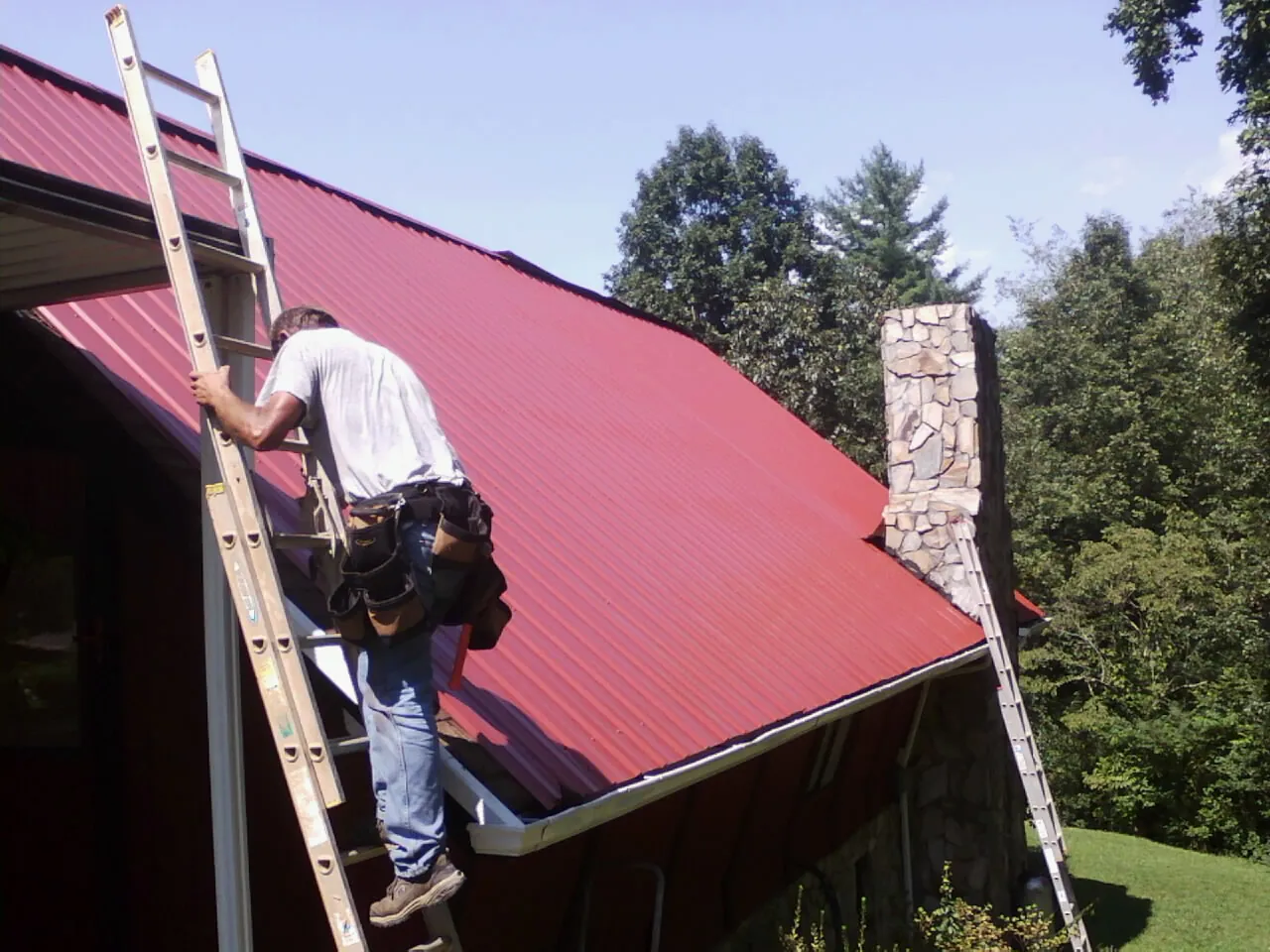 Roofer on a ladder beside a red metal roof and stone chimney — Soffit & Fascia Repair by Roofing Brothers in Hot Springs Village
